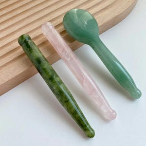 May include: Three gemstone facial tools: a green jade roller, a pink rose quartz roller, and a green jade spoon-shaped tool. The tools are arranged on a light wooden surface.