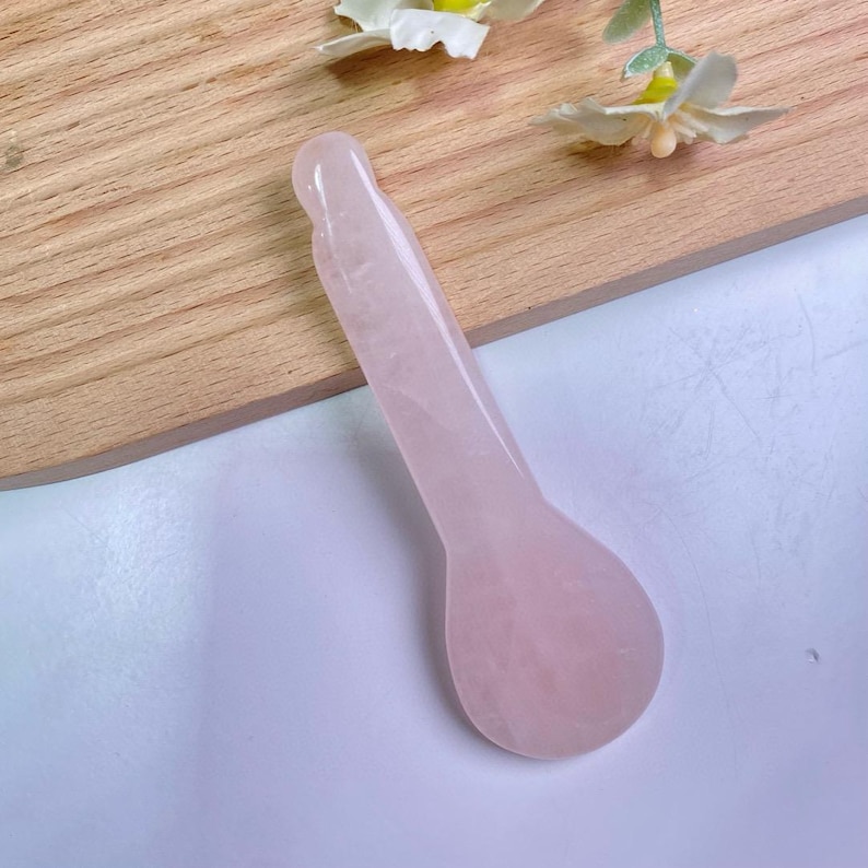May include: A smooth, pale pink rose quartz facial massage tool with a long, slender handle and a rounded, spoon-shaped end. The stone has a translucent, polished finish with subtle natural variations in its light pink color and texture.