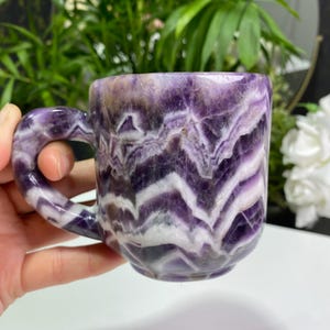 Może przedstawiać: Fioletowy kubek z ametystu z białym wzorem w postaci zwojów. Kubek ma uchwyt i jest wykonany z naturalnego kamienia.