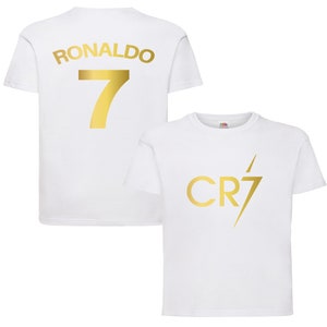 Pode incluir: Camiseta branca com um "RONALDO 7" dourado nas costas e um "CR7" dourado com um raio na frente.
