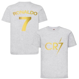 Pode incluir: Duas camisas cinzas com letras douradas. Uma camisa tem "RONALDO 7" nas costas. A outra camisa tem "CR7" com um raio.