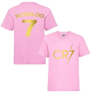Pode incluir: Camiseta rosa com um "RONALDO 7" dourado nas costas e um logotipo "CR7" dourado na frente.
