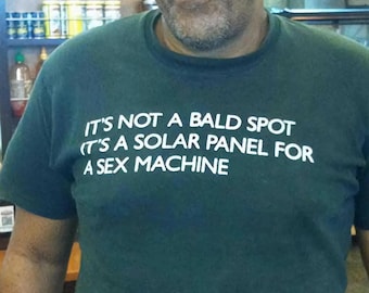 May include: Dark green t-shirt with the text "IT'S NOT A BALD SPOT (IT'S A SOLAR PANEL FOR A SEX MACHINE)" in white. The shirt is short-sleeved and has some white stains on the front.