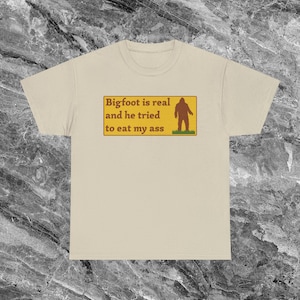 Könnte beinhalten: Beigefarbenes T-Shirt mit einem rechteckigen Aufdruck. Der Aufdruck zeigt den Text "Bigfoot is real and he tried to eat my ass" und eine braune Bigfoot-Silhouette auf grünem Gras.