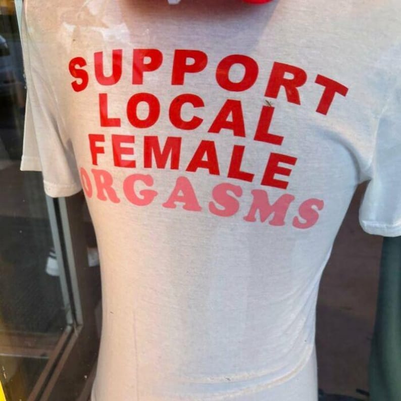 May include: White t-shirt with the text "SUPPORT LOCAL FEMALE ORGASMS" printed in red and pink.