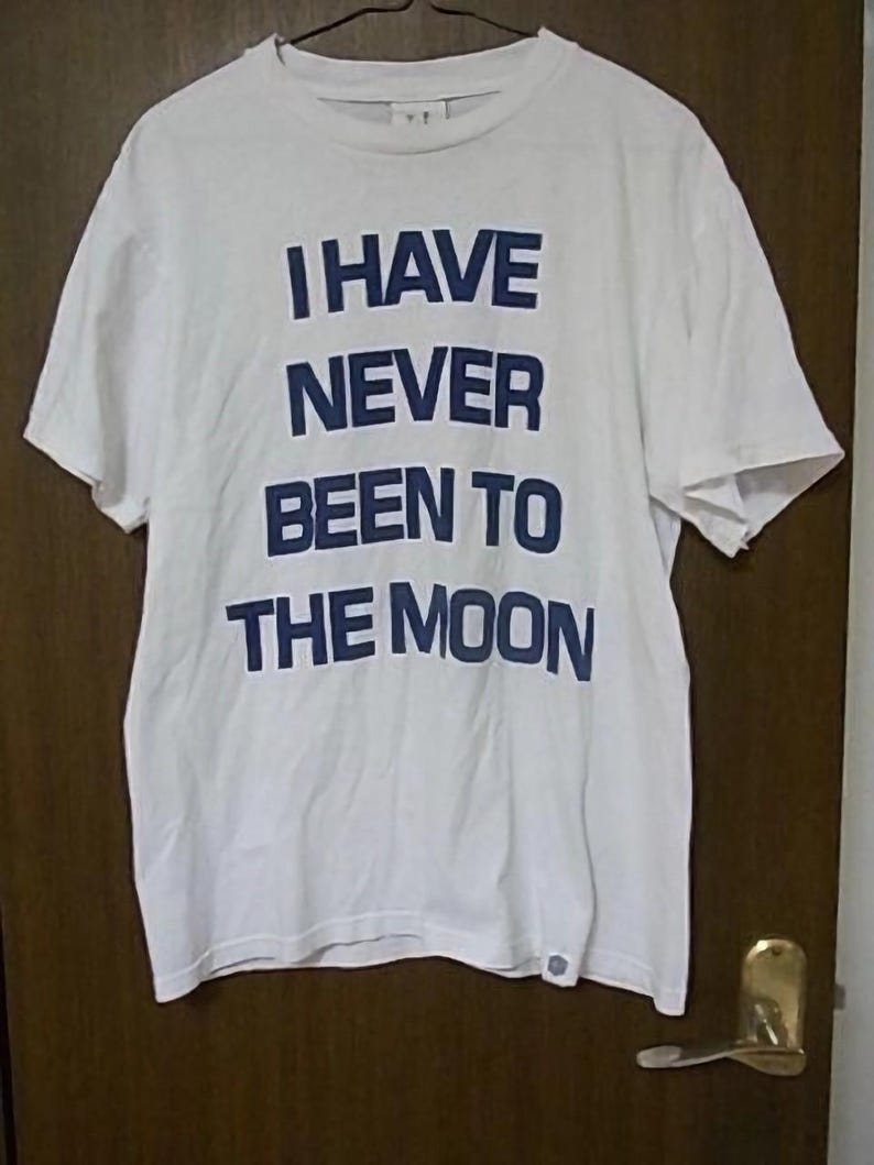 K&ouml;nnte beinhalten: Wei&szlig;es T-Shirt mit blauem Text, der "I HAVE NEVER BEEN TO THE MOON" lautet.
