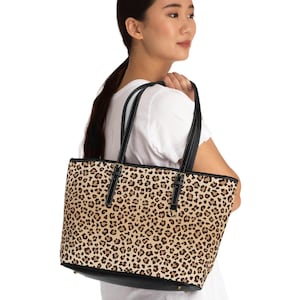 May include: A brown and black leopard print tote bag with black handles. The bag is being held by a person wearing a white t-shirt and black pants.