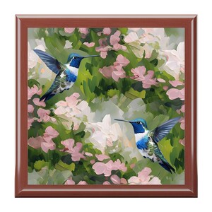 May include: A square wooden tray with a painting of two hummingbirds in flight. The birds have blue, white, and green feathers, with long beaks. The background features pink and white flowers and green foliage.