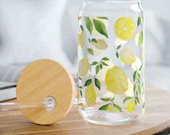 May include: A clear glass drinking cup with a bamboo lid and straw. The cup is decorated with yellow lemons and green leaves. The cup is sitting on a wooden table.