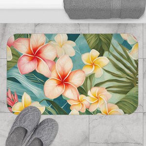 May include: A rectangular bath mat featuring a watercolor floral design. The design includes large pink and yellow plumeria flowers, green leaves, and a teal background. Gray slippers are at the bottom of the mat.