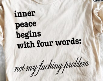 Puede incluir: Una camiseta de manga corta color crema con texto negro que dice: inner peace begins with four words: not my fucking problem. La prenda tiene cuello redondo y un corte estándar, mostrando la etiqueta de la marca Comfort Colors.