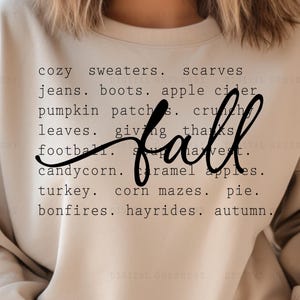 May include: A beige crewneck sweatshirt featuring black text that lists fall-themed words including cozy sweaters, scarves, jeans, boots, apple cider, pumpkin patches, crunchy leaves, giving thanks, football, harvest, candy corn, caramel apples, turkey, corn mazes, pie, bonfires, hayrides, and autumn, with the word fall written in a large script font.