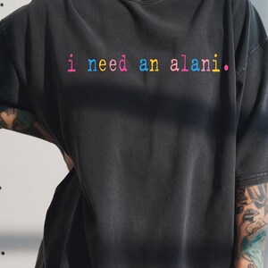 May include: Dark gray t-shirt with the phrase "i need an alani." in rainbow-colored text. The shirt has a relaxed fit and short sleeves. The text is in a playful, colorful font.