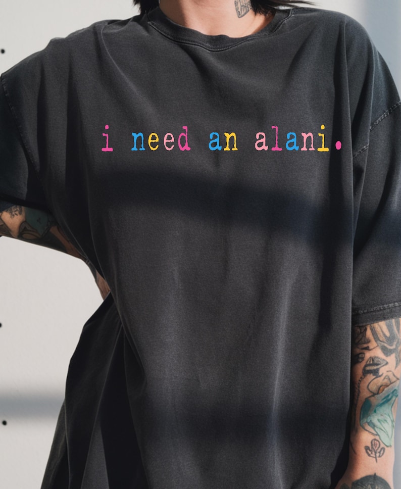 May include: Dark gray t-shirt with the phrase "i need an alani." in rainbow-colored text. The shirt has a relaxed fit and short sleeves. The text is in a playful, colorful font.