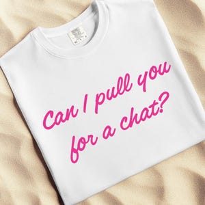 May include: White t-shirt with the text "Can I pull you for a chat?" in bright pink cursive lettering. The shirt is made of a soft material and is displayed on a sandy surface.