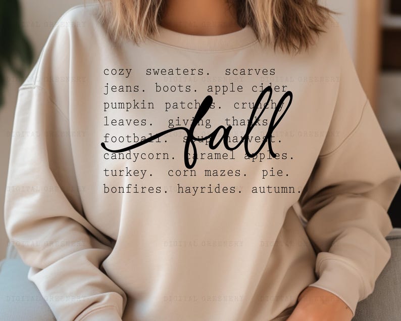 May include: A beige sweatshirt with the word "fall" in black text surrounded by a list of fall-themed words, including "cozy sweaters", "scarves", "jeans", "boots", "apple cider", "pumpkin patches", "crunch", "leaves", "giving thanks", "football", "candycorn", "caramel apples", "turkey", "corn mazes", "pie", "bonfires", "hayrides", and "autumn".