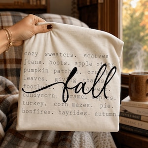 Pode incluir: Uma t-shirt cor de creme com texto preto listando palavras com tema de outono, incluindo cozy sweaters, scarves, jeans, boots, apple cider, pumpkin patches, crunchy leaves, giving thanks, football, soup, harvest, candycorn, caramel apples, turkey, corn mazes, pie, bonfires, hayrides, autumn, e a palavra fall em letra cursiva grande.