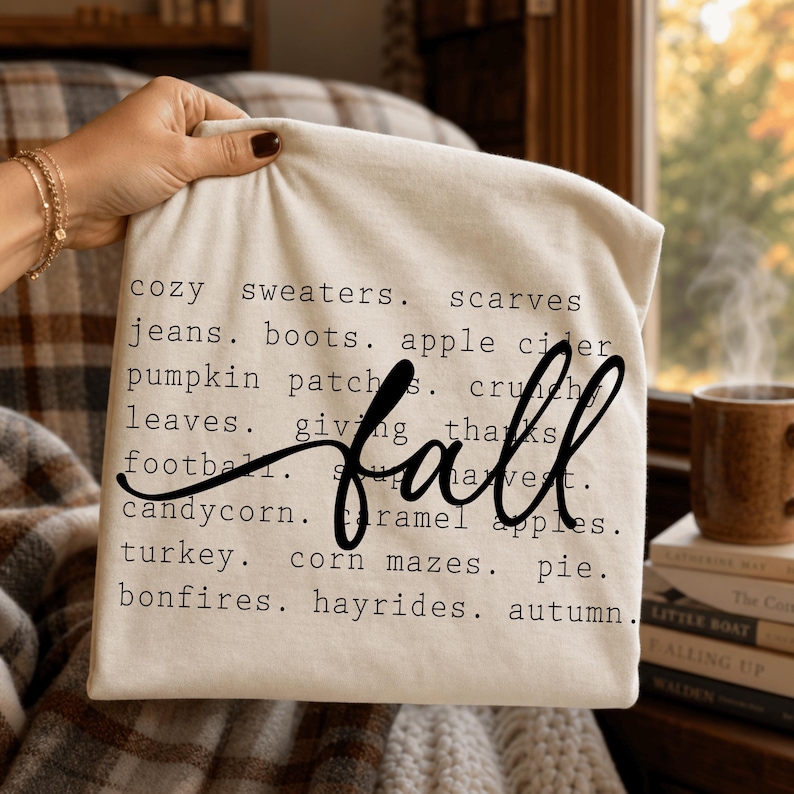 Pode incluir: Uma t-shirt cor de creme com texto preto listando palavras com tema de outono, incluindo cozy sweaters, scarves, jeans, boots, apple cider, pumpkin patches, crunchy leaves, giving thanks, football, soup, harvest, candycorn, caramel apples, turkey, corn mazes, pie, bonfires, hayrides, autumn, e a palavra fall em letra cursiva grande.