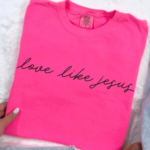 Puede incluir: Una camiseta rosa brillante con la frase "love like jesus" en cursiva. La camiseta tiene cuello redondo y está hecha de una tela suave. La etiqueta Comfort Colors es visible en la parte superior.