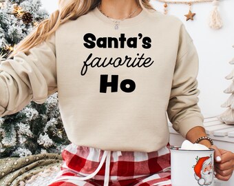 May include: A beige sweatshirt with the text "Santa's favorite Ho" printed in black. The sweatshirt is worn by a person wearing red and white plaid pajama pants.