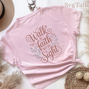 Può includere: T-shirt rosa chiaro con la scritta "Walk by faith not by sight" in marrone. Il design include elementi floreali e croci. È incluso anche il testo "2 Corinthians 5:7".