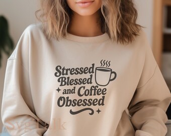 Puede incluir: Una sudadera beige con el texto "Stressed Blessed and Coffee Obsessed" en negro. El texto está en una fuente retro e incluye una taza de café estilizada con vapor saliendo de ella.