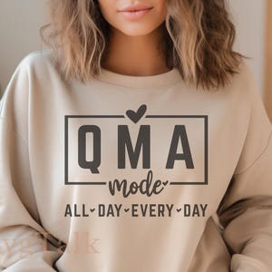 May include: A beige sweatshirt with the text "QMA mode" inside a black rectangle, with a heart above the "A". Below the rectangle is the text "ALL DAY EVERY DAY".