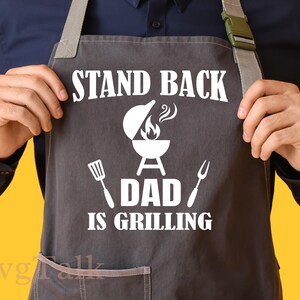 Może przedstawiać: Szary fartuch z białym tekstem i grafiką. Tekst brzmi "STAND BACK DAD IS GRILLING" nad grafiką grilla, szpatułką i widelcem. Fartuch ma kieszeń i szare paski.