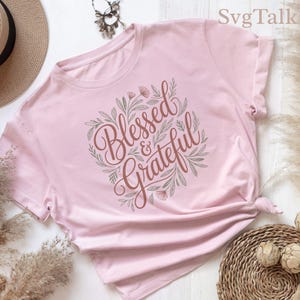Può includere: Una maglietta rosa chiaro con le parole "Blessed & Grateful" in un carattere marrone, circondata da una ghirlanda di foglie verdi e piccoli fiori rosa. La maglietta è annodata sul lato.