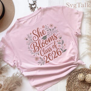 May include: A light pink t-shirt featuring a graphic print with the text She Blooms Class of 2026, a small graduation cap icon, and surrounding floral illustrations in shades of brown, green, and pink, centered on the front of the garment.