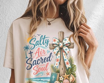 May include: A beige t-shirt featuring a graphic print with the text Salty Air Sacred Soul. The design includes a wooden cross with a floral bow, a small blue book with a cross, seashells, a starfish, palm trees, and a beach scene.