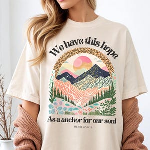 May include: Cream-colored t-shirt with the text "We have this hope" above a colorful mountain scene. The design includes a sun, mountains, and the text "As a anchor for our soul - HEBREWS 6:19". The shirt has short sleeves.