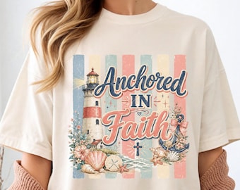 May include: Cream-colored t-shirt featuring the phrase "Anchored in Faith" in a decorative font. The design includes a lighthouse, anchor, seashells, and a cross, set against a backdrop of pastel stripes. The shirt has short sleeves.