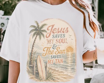 May include: A white short-sleeved t-shirt featuring a graphic of a beach scene with a palm tree, a surfboard, and a sunset. The text reads: Jesus SAVES MY SOUL & The Sun SAVES MY TAN. The design is enclosed in a soft-edged, oval-shaped frame.