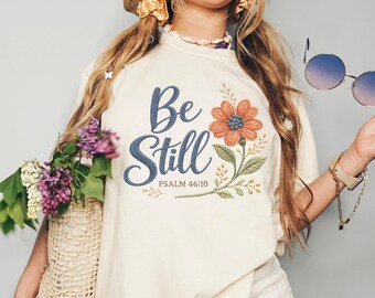 May include: An off-white short-sleeve t-shirt featuring the embroidered text Be Still and PSALM 46:10 in dark blue script, accompanied by a large orange flower with green leaves and stems, worn with white denim shorts and a straw bag.