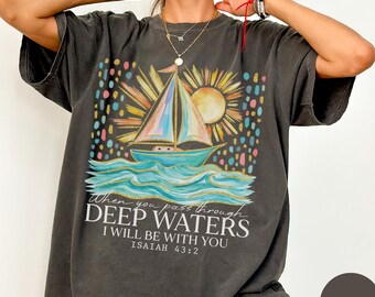 May include: Pepper-colored t-shirt with a watercolor design of a sailboat on the ocean, a sun, and the text "When you pass through deep waters I will be with you Isaiah 43:2." The shirt is part of the Comfort Colors collection.