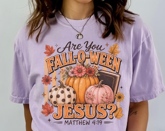 May include: A light purple short-sleeve t-shirt featuring a graphic print with the text Are You FALL-O-WEEN JESUS? MATTHEW 4:19. The design includes three pumpkins, one with polka dots, one with floral accents, and one topped with flowers, plus autumn leaves.