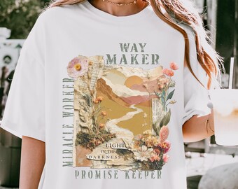 May include: White t-shirt with a graphic design featuring a landscape with a river, mountains, and flowers. The text on the shirt includes "WAY MAKER", "MIRACLE WORKER", "LIGHT IN THE DARKNESS", and "PROMISE KEEPER".