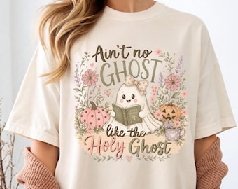 May include: A cream-colored t-shirt featuring a graphic of a cartoon ghost wearing a bow, holding a book labeled Holy Bible. The design includes pink pumpkins, flowers, and the text Ain't no GHOST like the Holy Ghost in brown and pink script.