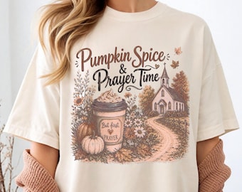 May include: A cream-colored t-shirt featuring a graphic print with the text "Pumpkin Spice & Prayer Time" and "But first PRAYER". The design includes a coffee cup, pumpkins, autumn foliage, and a church building along a dirt path.
