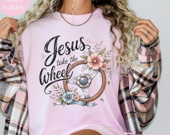 May include: A light pink t-shirt featuring a graphic of a steering wheel adorned with pink and blue flowers. The text "Jesus take the Wheel" is printed in a black script font above the steering wheel. The shirt is worn under a plaid flannel shirt.