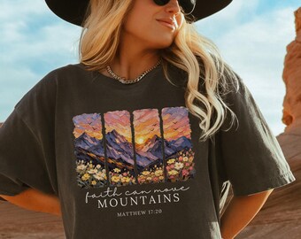 May include: Dark gray t-shirt featuring a colorful mountain landscape design with a sunset and floral accents. The text "faith can move mountains" and "Matthew 17:20" are printed below the image. The shirt has a relaxed fit.