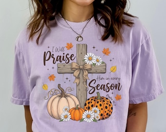 May include: A light purple t-shirt featuring a graphic of a wooden cross with a tan bow, surrounded by pumpkins, white daisies, and orange autumn leaves. The text reads I Will Praise Him in every Season and Psalm 46:2 in black script.