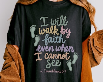 May include: A black t-shirt featuring the text "I will walk by faith even when I cannot see" in multi-colored script lettering, accompanied by small green baby footprint graphics and the citation "2 Corinthians 5:7" printed in pink.