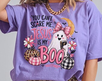 May include: A light purple short-sleeve t-shirt featuring a graphic print of a ghost wearing a pink plaid bow and holding a book. The text reads YOU CAN'T SCARE ME... JESUS IS MY BOO, surrounded by pink flowers, a crescent moon, and three pumpkins.