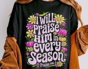 May include: A black t-shirt featuring a graphic print with the text "I WILL PRAISE HIM IN EVERY SEASON" and "Psalm 146:2" in a wavy font. The design is surrounded by colorful illustrations of yellow and pink flowers and small pumpkins.