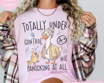 May include: A light pink t-shirt featuring a graphic of a goose wearing glasses and a cardigan, a small duckling, and the text "TOTALLY UNDER CONTROL NOT PANICKING AT ALL" with an "A+" grade symbol and the hashtag "#backtoschool" printed in black.