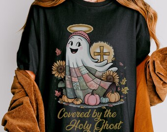 May include: A black t-shirt featuring a graphic of a ghost wearing a beanie and halo, draped in a patchwork quilt. The design includes sunflowers, pumpkins, a cross, and the text "Covered by the Holy Ghost" in a gold script font.