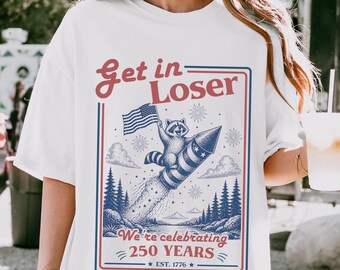 May include: White t-shirt with a patriotic graphic. The design features a raccoon riding a rocket with an American flag, fireworks, and the text "Get in Loser" and "We're celebrating 250 YEARS".