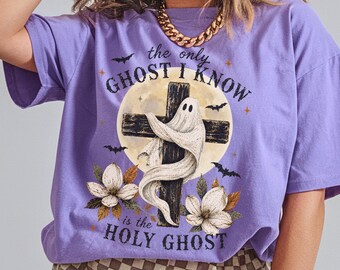 May include: A light purple short-sleeve t-shirt featuring a graphic print of a white ghost wrapped around a dark cross in front of a full moon. The text reads, "the only GHOST I KNOW is the HOLY GHOST," surrounded by bats and white flowers.